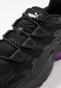 Svart atletisk sneaker med nätöverdel, texturerade syntetiska överlägg, vadderad krage och lila accenter på sulan. Reflekterande detaljer och snörning.