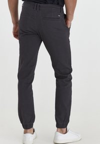 Harmaat puuvillaiset juoksuhousut, joissa on joustavat resorit, takataskut ja logopatchi. Yhdistettynä valkoisiin lenkkareihin neutraalilla taustalla.