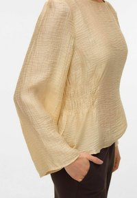 Blusa beige chiaro con tessuto testurizzato e arricciato, caratterizzata da maniche lunghe e vita arricciata per una vestibilità strutturata.