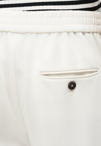 Κοντινό πλάνο λευκών παντελονιών με ελαστική μέση, πίσω τσέπη με σκούρο κουμπί και ριγέ κορυφαία ρίγα στην επάνω πλευρά.