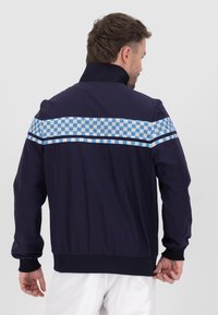 Giubbotto antivento navy con tessuto testurizzato, caratterizzato da una striscia orizzontale a scacchi blu chiaro sulla schiena e polsini a costine.