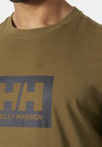 Primer plano de una persona que lleva una camiseta de cuello redondo de color verde oliva con un logo negro de Helly Hansen en el pecho.