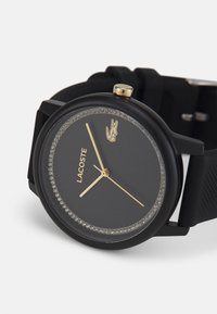 Juodas silikono laikrodis su apvaliu ciferblatu, tamsiu skaičiumi, auksiniais akcentais ir blizgančiu ratu. Yra įtrauktas Lacoste logotipas ir paprasti valandų žymenys.