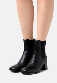 Černé kotníkové boty vyrobené z hladké kůže, s elastickými postranními panely a čtveratou špičkou. Podpatky jsou robustní a mírně vysoké.
