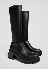 Bottes noires jusqu'aux genoux en cuir lisse. Dotées d'une semelle plate-forme épaisse et d'une fermeture éclair sur le côté pour un enfilage facile. Design épuré et moderne.