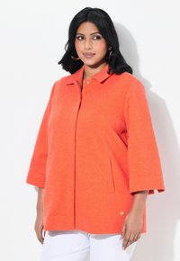 Oranžová strukturovaná bunda s límcem, třičtvrtinovými rukávy, bočními kapsami a zlatým akcentem blízko lemování.
