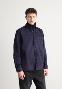 Cămașă din denim de culoare mov închis, cu guler cu nasturi, buzunar frontal și nasturi negri. Purtați peste un pulover cu guler înalt negru, asortat cu blugi negri.