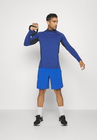 Blauw sportshirt met lange mouwen en zwarte accenten, voorzien van een hoge kraag, gecombineerd met heldere blauwe shorts. Het model houdt een kettlebell vast.