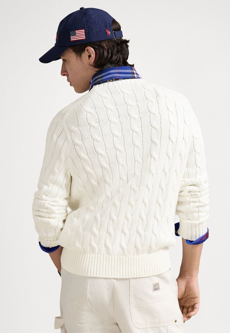 Vit kabelstickad tröja med ribbstickade muddar och fåll, bärs över en blå rutig skjorta, kombinerad med en marinblå keps med broderad logotyp.