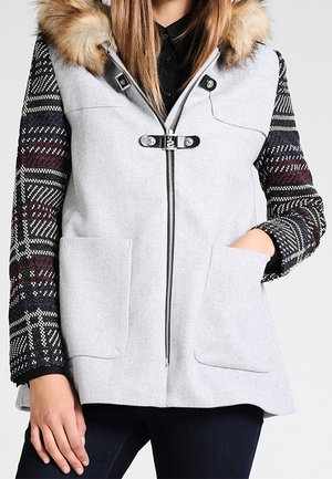 Femme portant un manteau sans manches gris clair avec de grandes poches, une capuche bordée de fourrure et des manches longues à motifs. Une main est dans une poche.