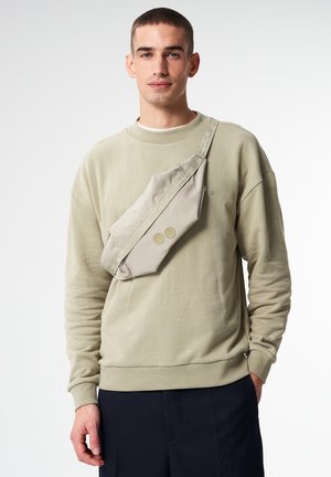 Lichtbeige crossbody tas van zacht stof, met twee ronde knopen en een verstelbare schouderband, gedragen over een gedempt groene sweatshirt.