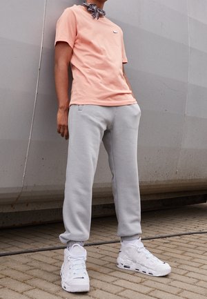 Jasnoróżowa bawełniana koszulka z małym logo, szare spodnie dresowe i białe buty sportowe. Zawiera czarno-biały, wzorzysty szalik na szyję.