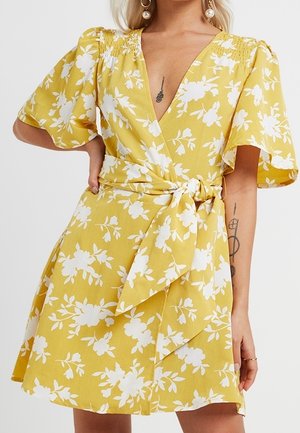 Żółta sukienka kopertowa z białym kwiatowym nadrukiem, krótkimi bufiastymi rękawami, głębokim dekoltem w kształcie litery V oraz przewiązanym paskiem w talii, noszona przez osobę z tatuażami na ramionach i perłowymi kolczykami.