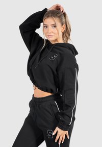 Schwarzer, kürzer geschnittener Hoodie mit Kordelzug, der mit einem gestickten "S"-Logo verziert ist. Kombiniert mit passenden schwarzen Jogginghosen und weißen Akzenten. Weicher Stoff.