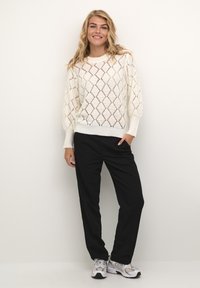 Weißer Strickpullover mit Diamantmuster, gerippten Bündchen und Saum, kombiniert mit schwarzen Hosen und grauen Sneakers. Glatte, luftige Textur.