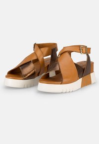 Sandales compensées en cuir marron avec design à lanières croisées, dotées d'un accent métallique et d'une semelle en caoutchouc blanc avec un profil texturé.