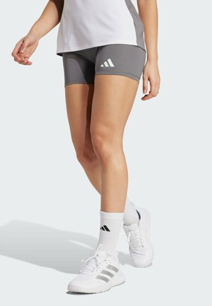 Personne portant un short de sport gris, des chaussettes blanches, des baskets blanches et un haut de sport blanc et gris, debout avec une jambe légèrement en avant.