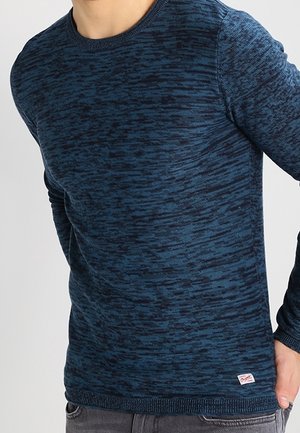 Mężczyzna w swetrze z długim rękawem i okrągłym dekoltem, w niebieski i ciemnoniebieski wzór strukturalny, stoi z jedną ręką w kieszeni.