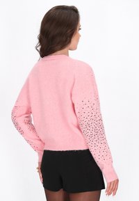 Frau mit langen braunen Haaren, die einen rosa Pullover mit schwarzen Pailletten an den Ärmeln und schwarze Shorts trägt, seitlich vor einem weißen Hintergrund stehend.