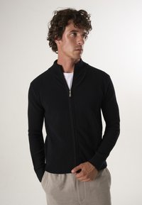 Zwarte, geribbelde zip-up jas met een hoge kraag, met lange mouwen en een ontspannen fit, gecombineerd met beige broeken.