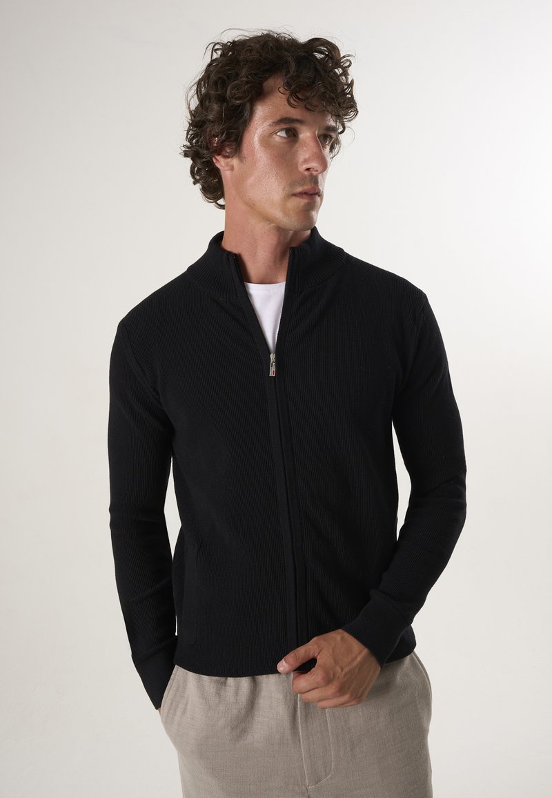 Zwarte, geribbelde zip-up jas met een hoge kraag, met lange mouwen en een ontspannen fit, gecombineerd met beige broeken.