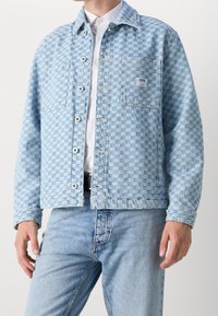 Light blue denim jacket featuring a checkerboard pattern, button-up front, two chest pockets, and a branded tag on the left pocket.
