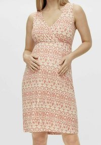 Mujer embarazada con un vestido de tirantes de patrón melocotón y crema, con las manos descansando suavemente sobre su vientre frente a un fondo sencillo.