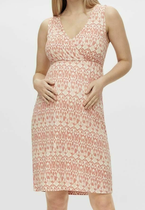 Mujer embarazada con un vestido de tirantes de patrón melocotón y crema, con las manos descansando suavemente sobre su vientre frente a un fondo sencillo.