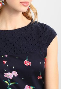 Blusa navy con stampa floreale, caratterizzata da un sovrapposto testurizzato con motivi a taglio, maniche a cappuccio e una finitura in tessuto liscia.