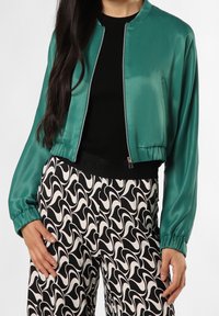 Satin bomberjakke i teal, med lynlås foran, ribbede ærmer, parret med bukser i sort og hvidt svirlemønster.