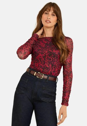 Langärmliges Oberteil in Rot mit schwarzem Tiermuster, aus durchsichtiger Stoff gefertigt. Kombiniert mit dunklen, hochtaillierten Jeans und einem braunen Ledergürtel.