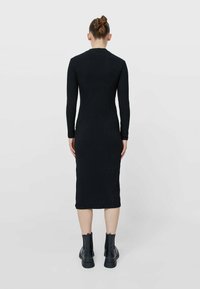 Robe en maille côtelée noire avec manches longues, silhouette ajustée, longueur au genou, et détail d'ourlet brut, associée à des bottines noires.