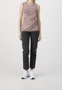 Ärmlös ribbad topp i dammig rosa, med djärva grafik och text i mörkare nyanser; kombinerad med svarta slim-fit jeans och vita sneakers.