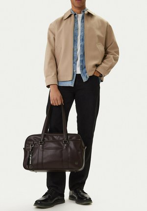 Uomo che indossa una giacca beige, camicia di jeans, maglietta bianca, pantaloni neri, scarpe nere, che tiene una borsa porta documenti in pelle marrone scuro davanti a uno sfondo uniforme.