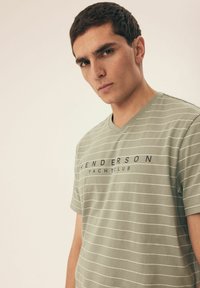 Zielona w paski koszulka z bawełny, z dekoltem w kształcie litery V, krótkimi rękawami i centralnie nadrukowanym logo w ciemnoniebieskim kolorze z tekstem.