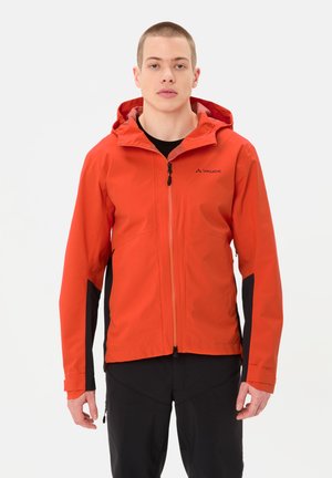 Jonge man draagt een feloranje capuchonjas met zwarte zijpanelen en een zwarte broek, staand tegen een effen achtergrond.