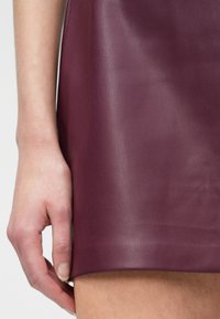 Rechterhand rustend op de dij, gekleed in een korte bordeauxleren rok, die de contrast tussen huidkleur en de textuur van het leer benadrukt.