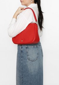 Borsa a spalla in pelle rossa con forma curva, finitura testurizzata e chiusura magnetica. I dettagli includono una cerniera argentata e accenti.