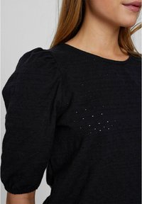 Haut noir texturé avec manches bouffantes, encolure ronde et perforations décoratives sur le tissu, créant un détail subtil.