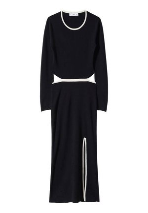 Femme portant une robe noire ajustée à manches longues, bordure blanche, découpe latérale à la taille et fente jusqu'à la cuisse, posant avec les mains sur les hanches.