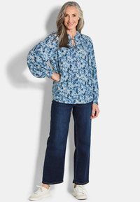 Blaue Bluse mit floralen Muster und gebundenem Kragen, lockeren Ärmeln, kombiniert mit dunkelblauen geraden Jeans und weißen Sneakern.