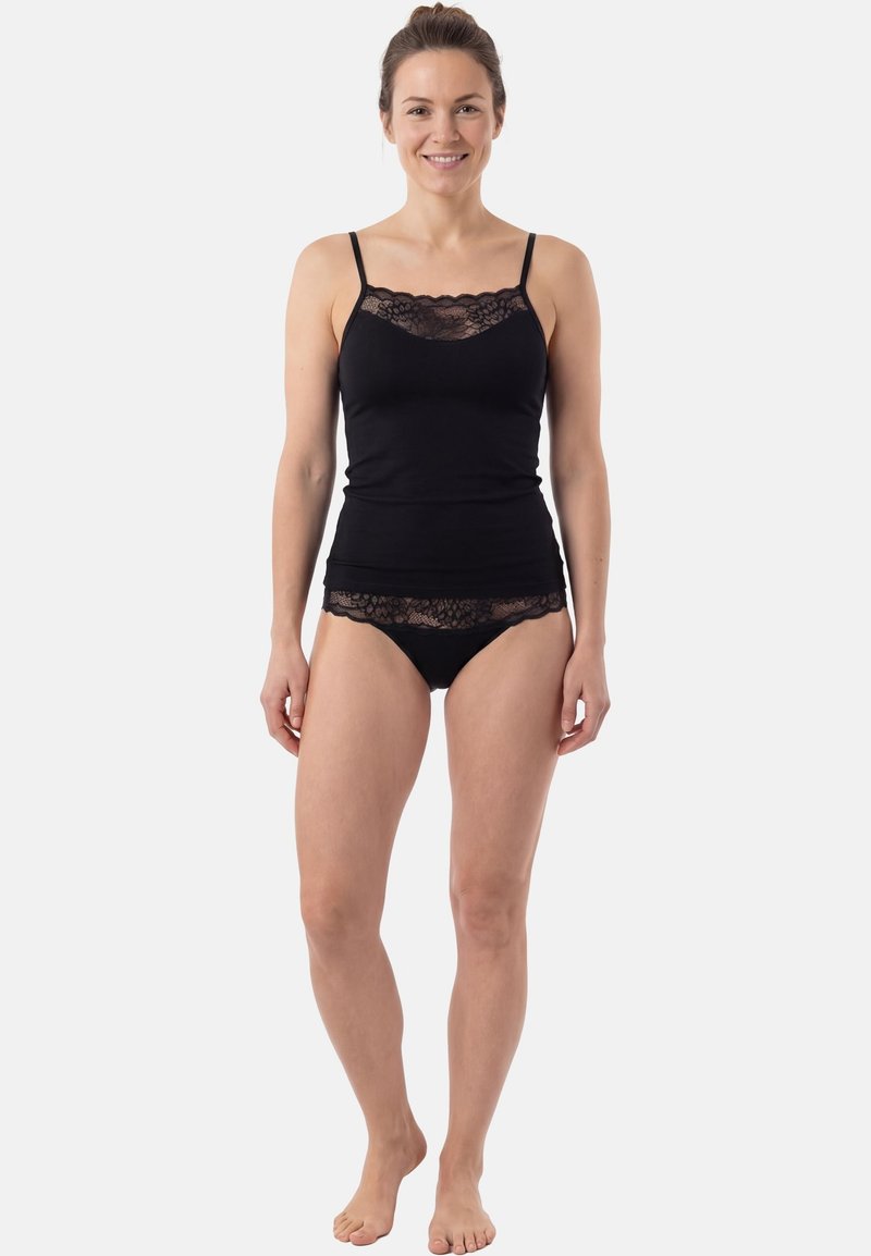 Femme debout pieds nus portant un caraco noir orné de dentelle et une lingerie noire assortie avec bordure en dentelle, sur un fond clair uni.