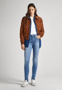 Chaqueta bomber de ante marrón con bordado floral, combinada con una camisa de denim azul. Jeans skinny azul claro y zapatillas blancas completan el look.
