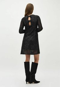 Robe noire à sequins avec manches longues, encolure ronde et détail dos en forme de trou de serrure. Associée à des bottes en daim noir montantes jusqu'au genou.