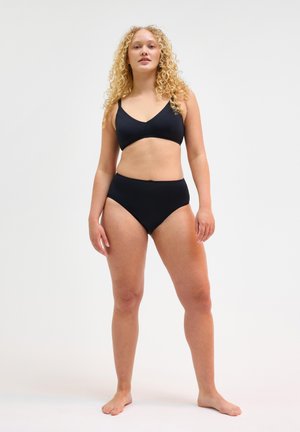 Giovane donna con capelli biondi ricci che indossa reggiseno nero e biancheria a vita alta, in piedi scalza su uno sfondo chiaro uniforme.