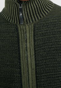 Nahaufnahme eines grün-schwarzen Strickpullovers mit einem hochgerippten Kragen und einem teilweise heruntergezogenen schwarzen Reißverschluss vorne.