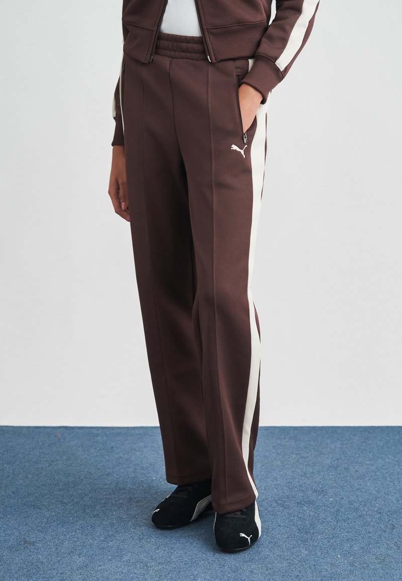 Καφέ αθλητικό παντελόνι με λευκές ρίγες στο πλάι, ελαστική μέση και τσέπες με φερμουάρ. Κατασκευασμένο από υφασμάτινη υφή, συνδυασμένο με μαύρα αθλητικά παπούτσια.