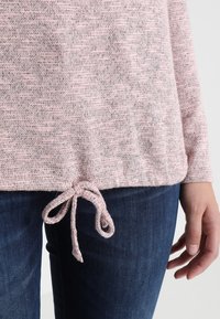 Top lavorato a maglia rosa chiaro con un motivo tessuto, dotato di orlo con coulisse e maniche lunghe, abbinato a jeans blu scuro.