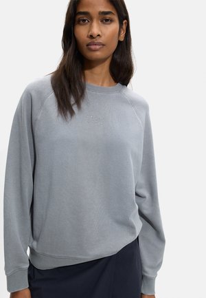 Kobieta nosząca luźną, jasnoszarą bluzę z małym haftowanym napisem na piersi, zestawioną z ciemną spódnicą na jednolitym tle.