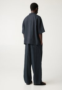 Set di camicia e pantaloni in lino scuri e larghi. La camicia presenta un colletto e maniche corte; i pantaloni hanno un taglio ampio con tasche laterali.